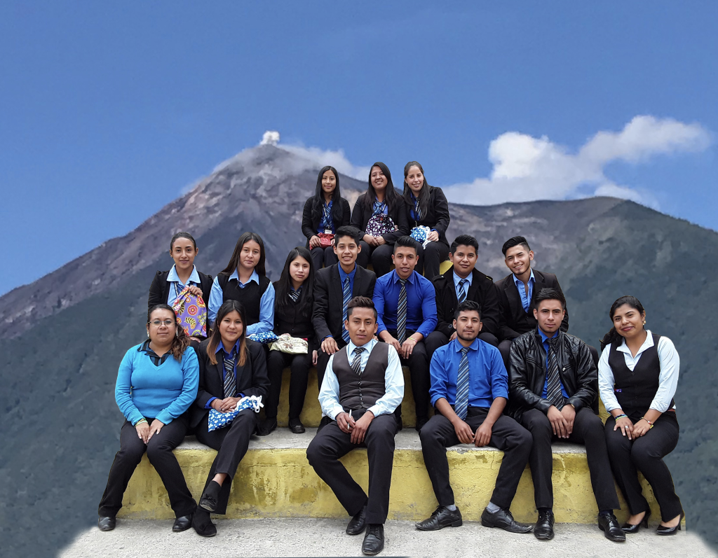 About 20 nicely dressed teenage kids sitting on a stair pyramid posing for a photo with a volcano in the background
